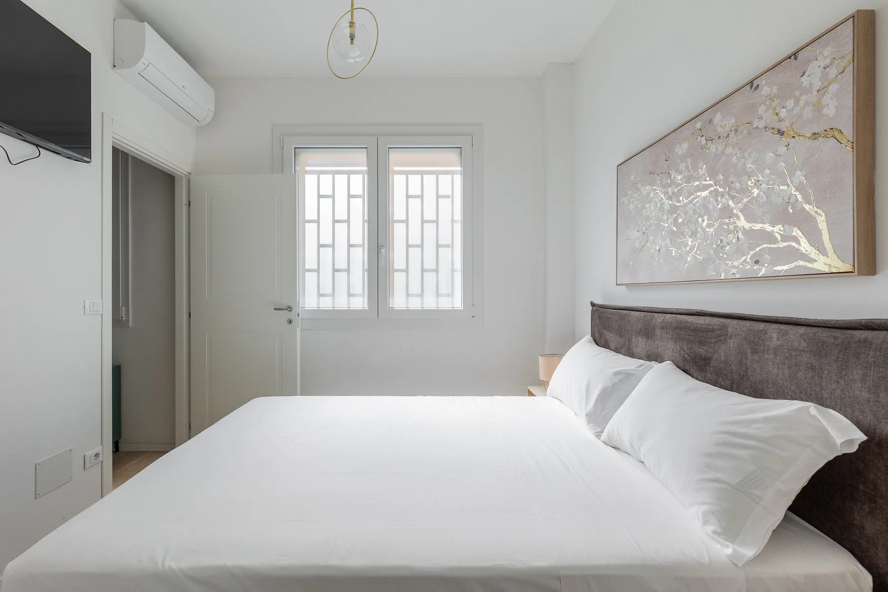 A bright bedroom with a neatly made bed beneath an abstract wall painting.