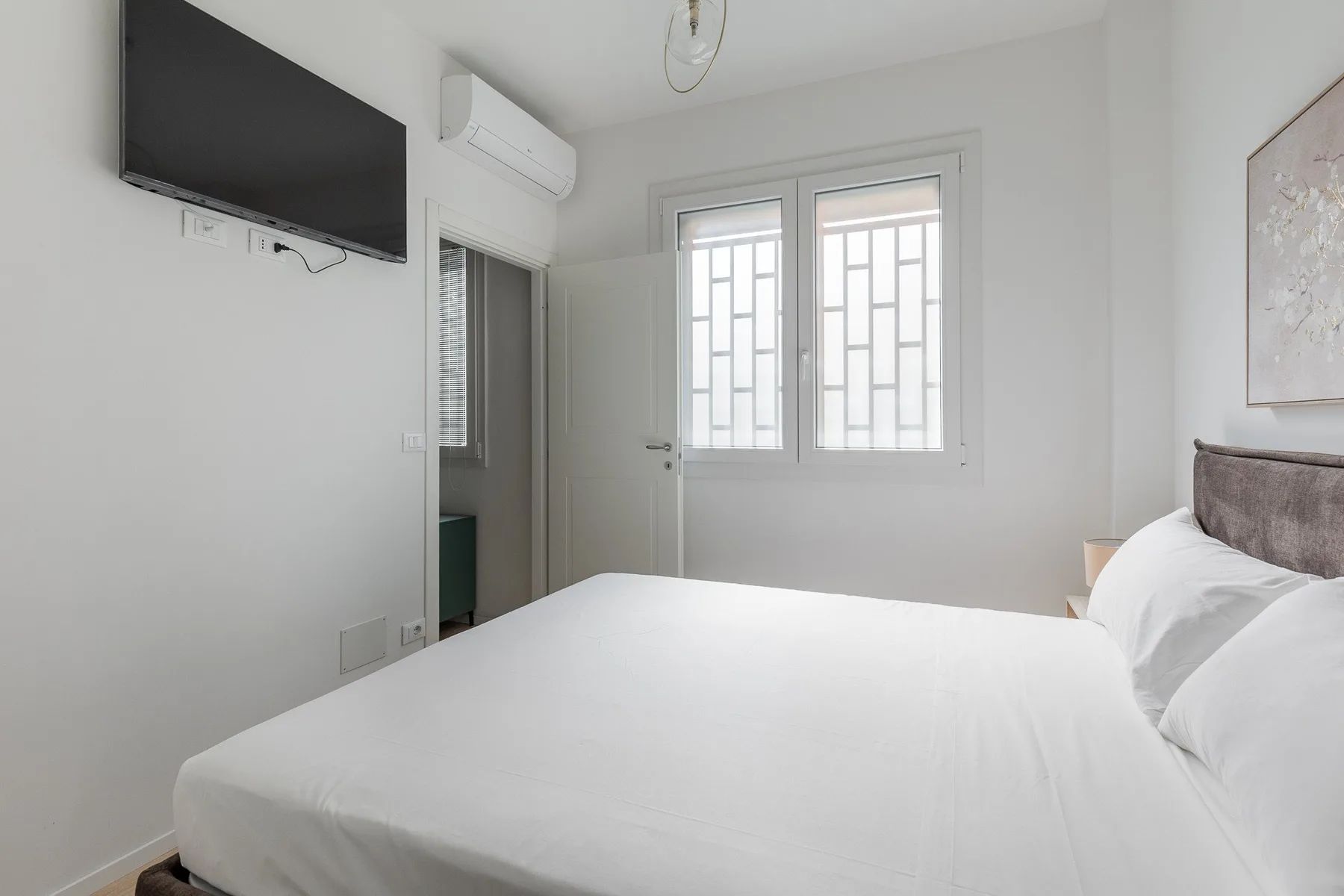 A bright bedroom with a bed beneath two windows and a wall-mounted television.