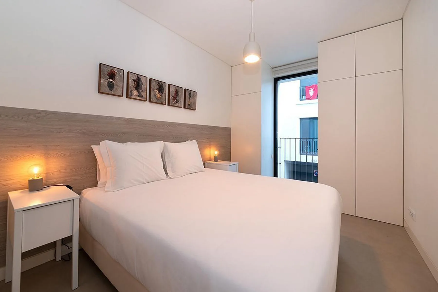 Un dormitorio con cama doble, mesillas de noche y una ventana con vistas a edificios cercanos.