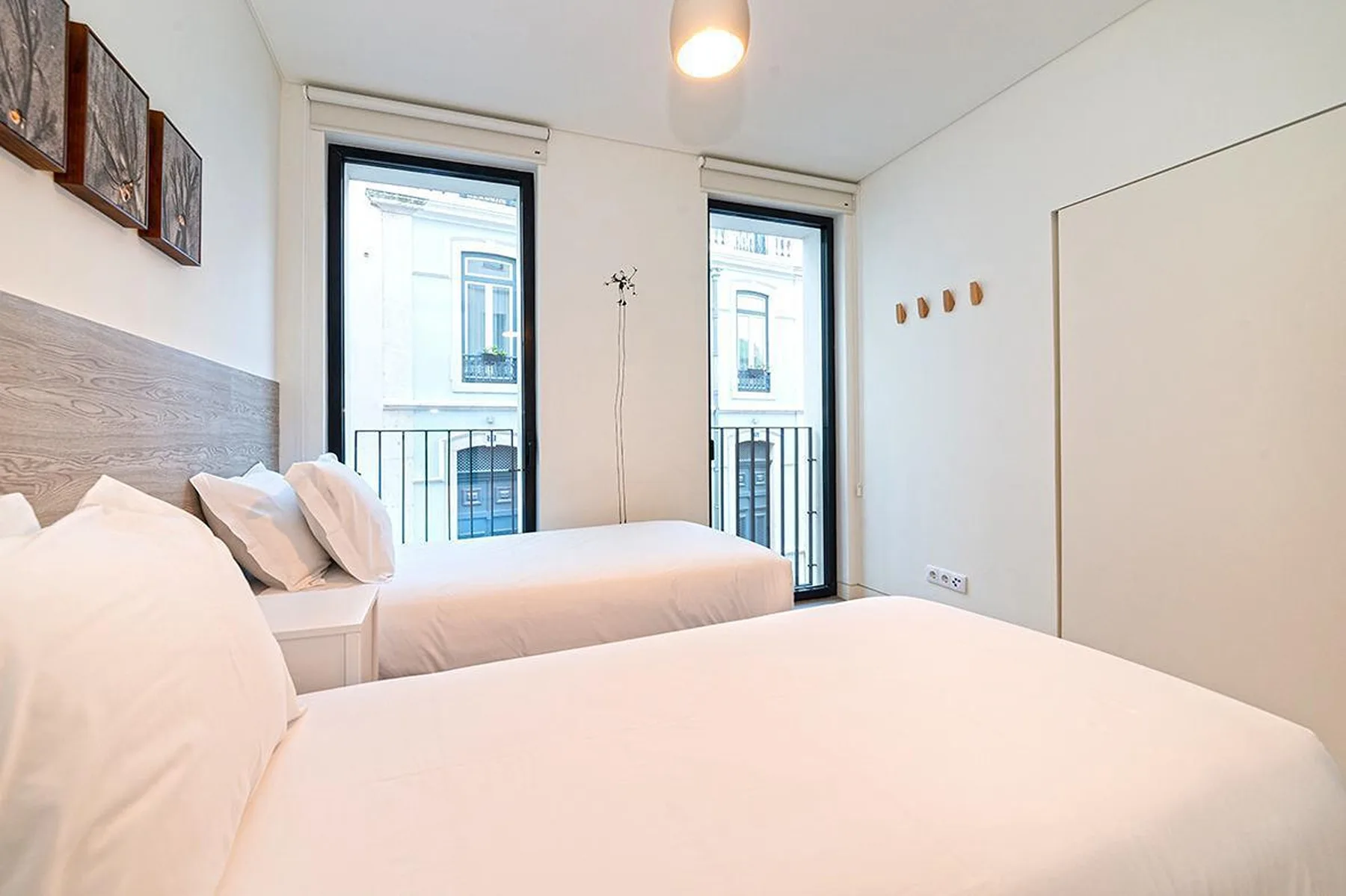 Un dormitorio con dos camas y ventanas altas frente a un edificio cercano.