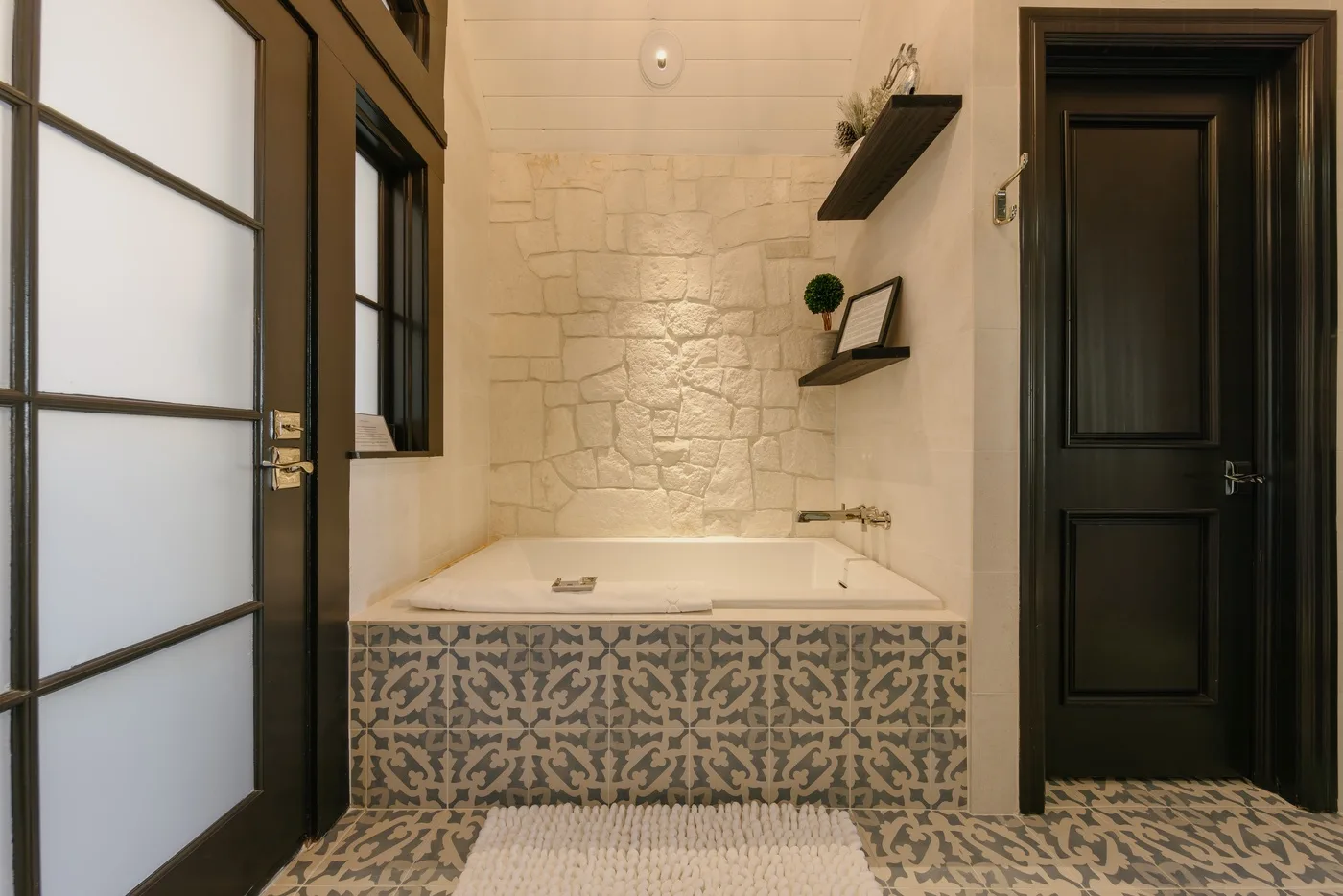 Un baño con bañera empotrada, azulejos estampados y una pared de piedra clara.