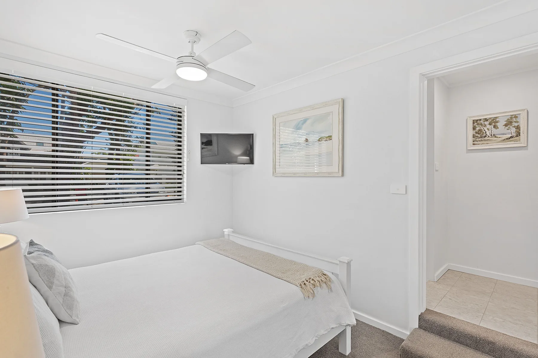 A bright bedroom with a bed, window blinds, and a wall-mounted television.