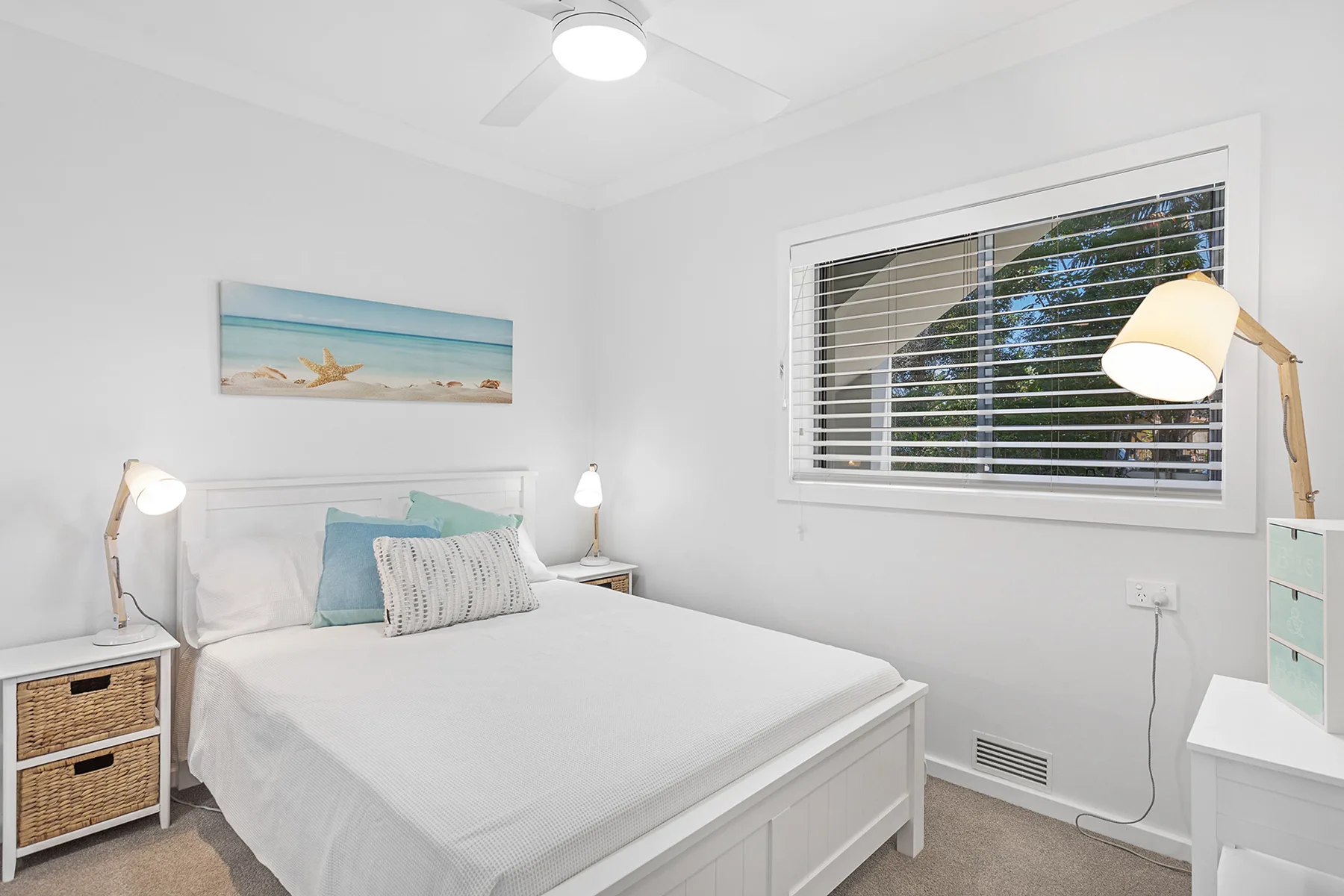 A bright bedroom with a neatly made bed beneath a window with blinds.