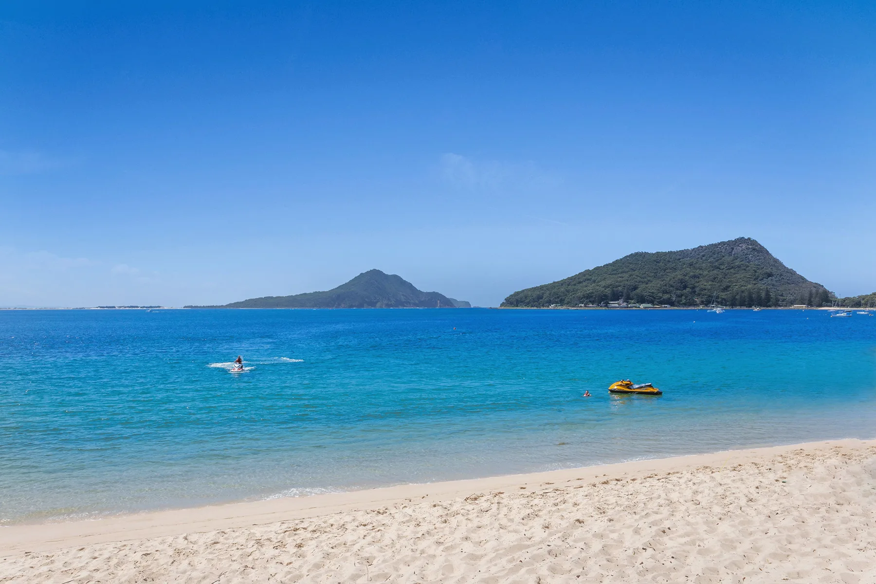 A sandy beach meets clear blue water, with green islands visible across the bay.