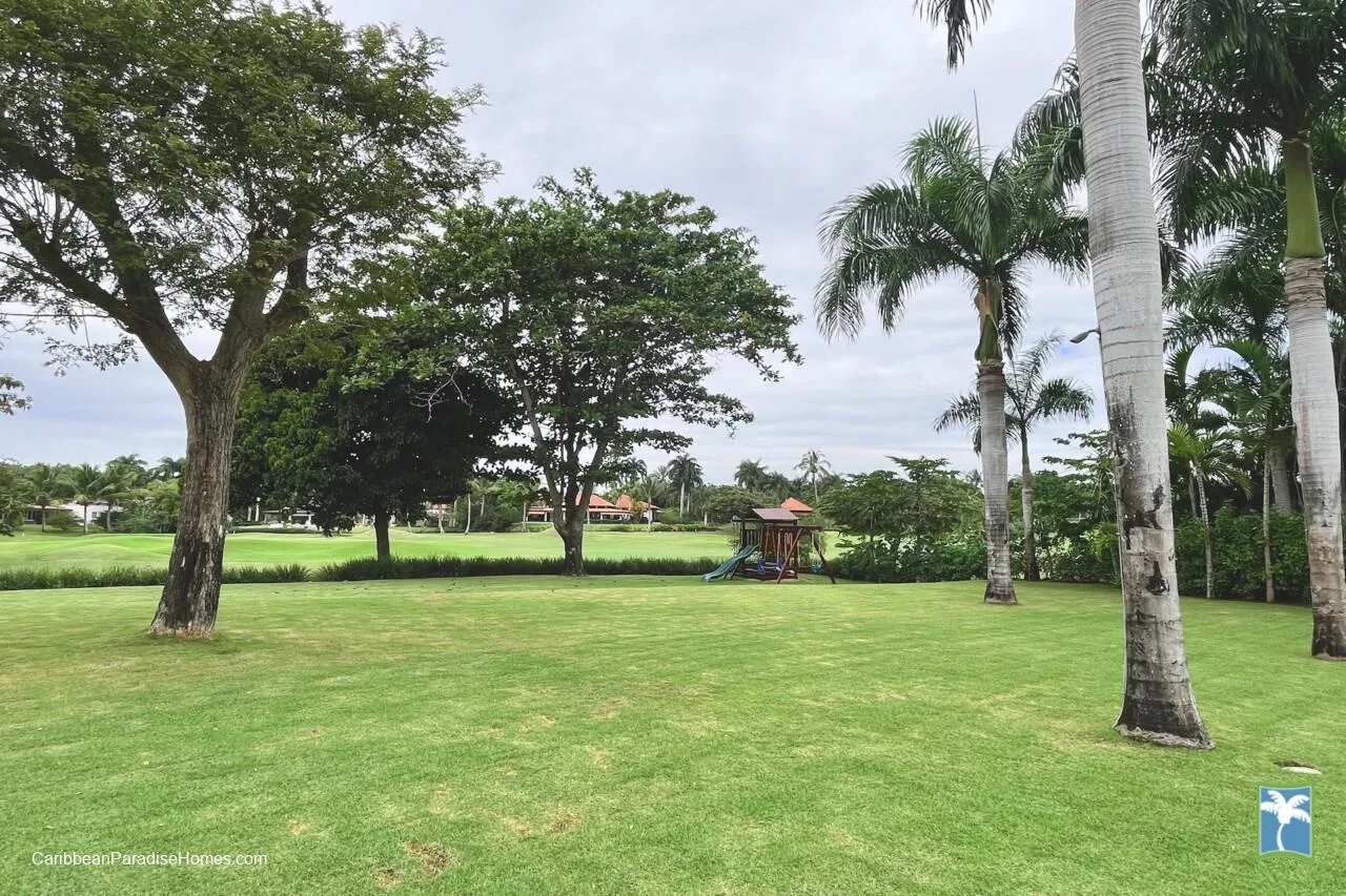 Un parque con césped, árboles maduros y un pequeño parque infantil a lo lejos.