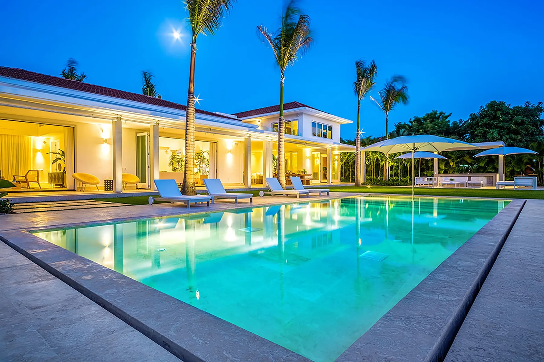 Una casa moderna iluminada da a una piscina al anochecer, con palmeras cerca.