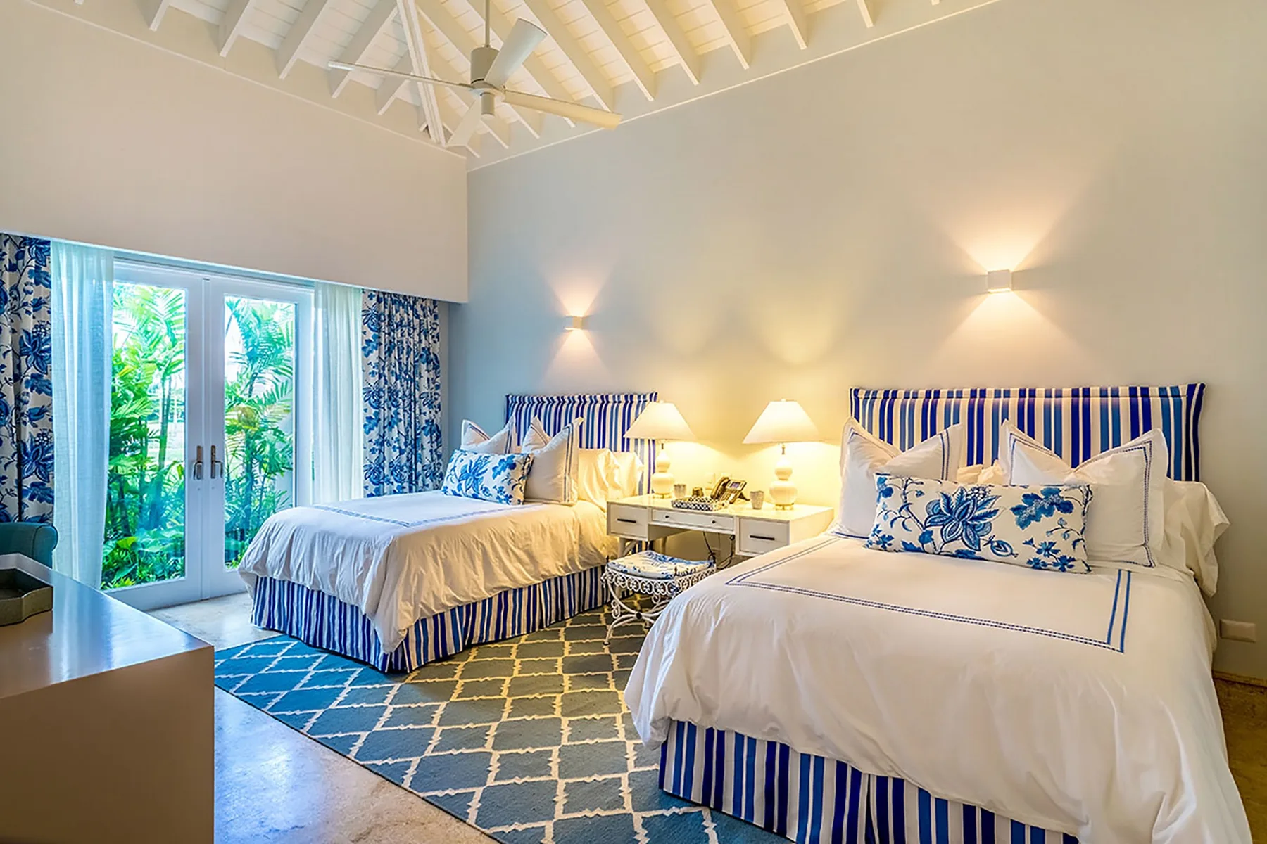 Un dormitorio luminoso con dos camas, detalles azules y puertas de vidrio junto a la vegetación.