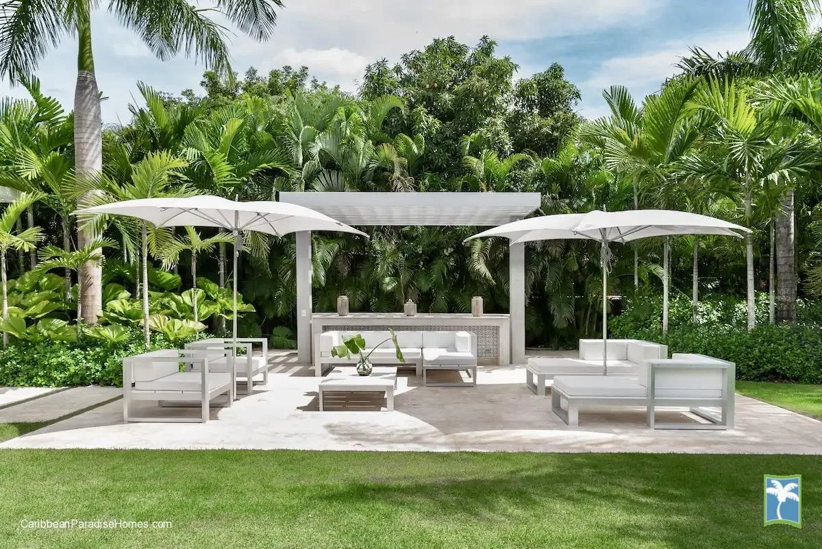 Una zona de estar al aire libre con asientos blancos y sombrillas entre vegetación tropical.