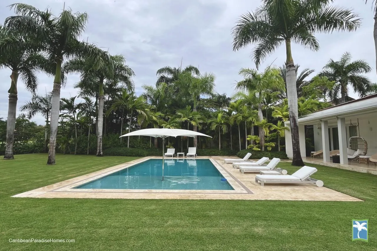 Una piscina está junto al césped, con tumbonas y palmeras cerca.