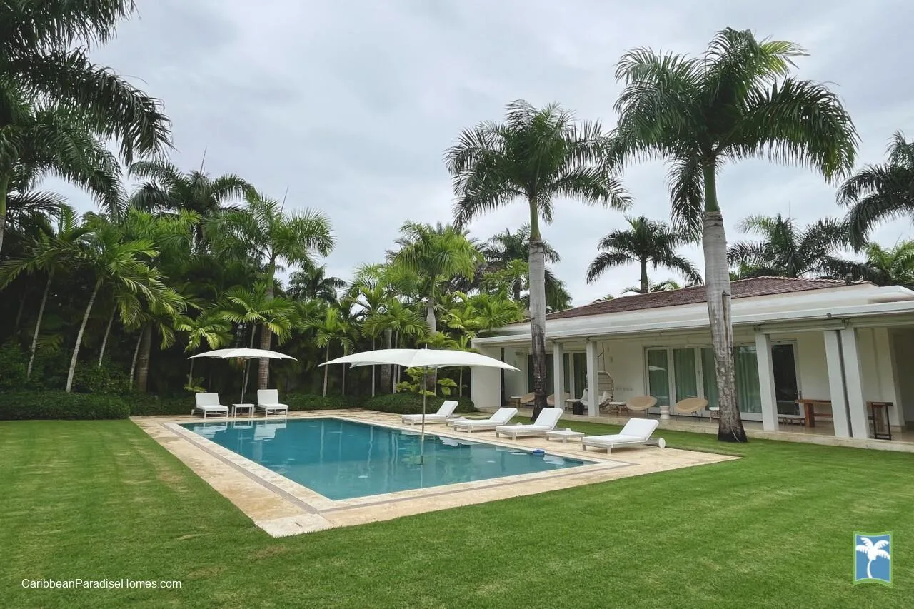 Una piscina al aire libre junto a un edificio blanco, rodeada de palmeras y césped.