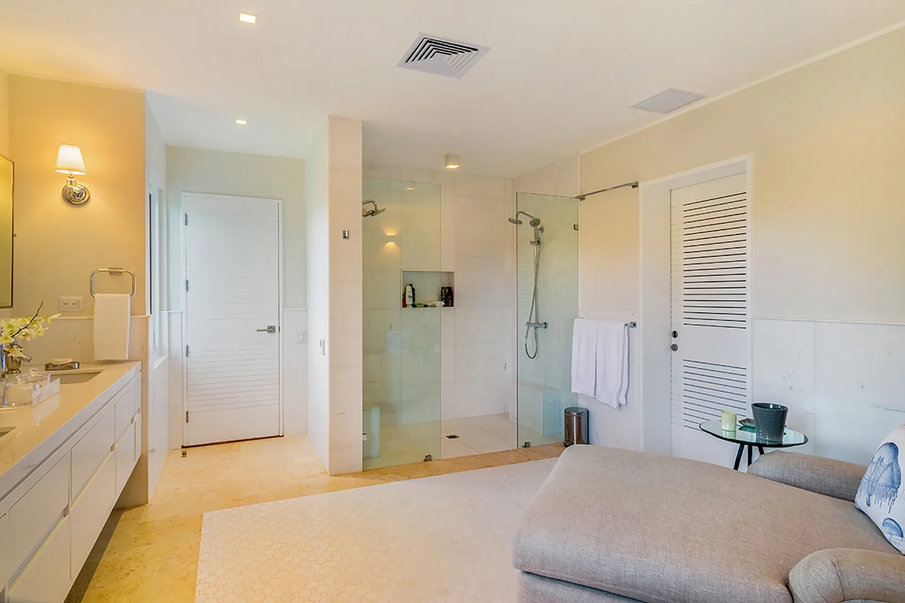 Un baño luminoso con lavabo, ducha de vidrio y asiento tapizado.
