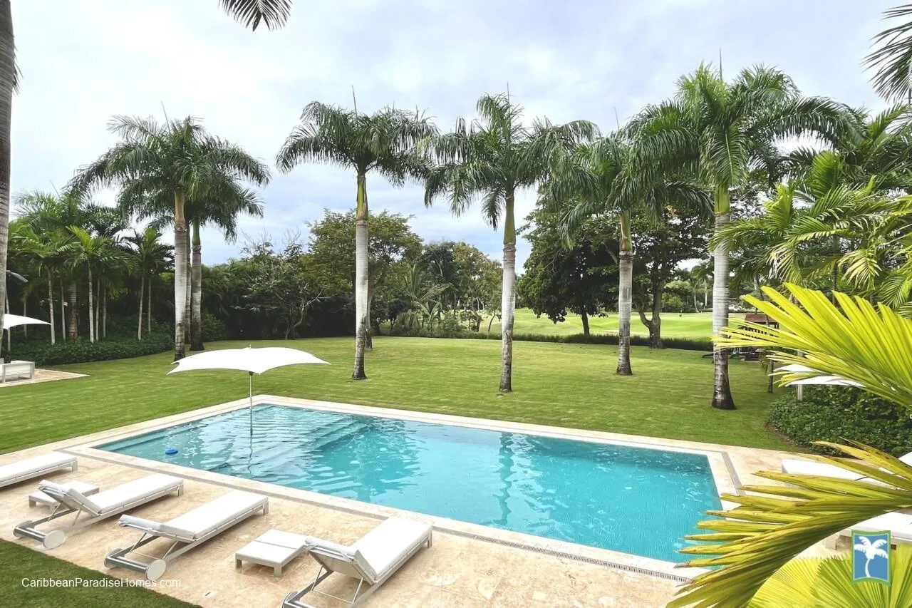 Una piscina junto a un césped, con palmeras y asientos para descansar.
