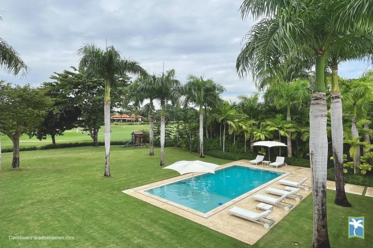 Una piscina al aire libre está rodeada de césped ajardinado, palmeras y tumbonas.