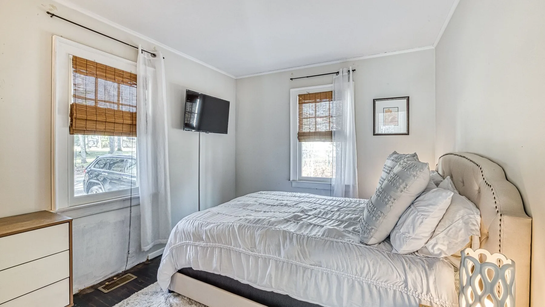 Un dormitorio luminoso con una cama, dos ventanas y un televisor montado en la pared.