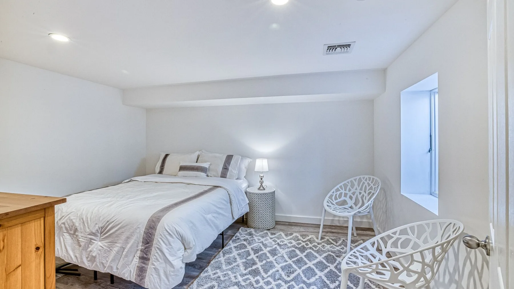 Un dormitorio luminoso con una cama tendida, una alfombra estampada y asientos blancos.