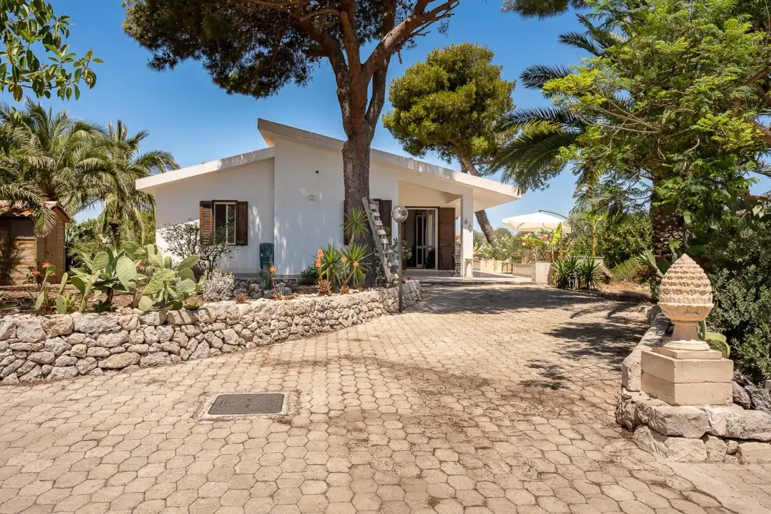 Una casa blanca está junto a un patio pavimentado y sombreado por árboles.