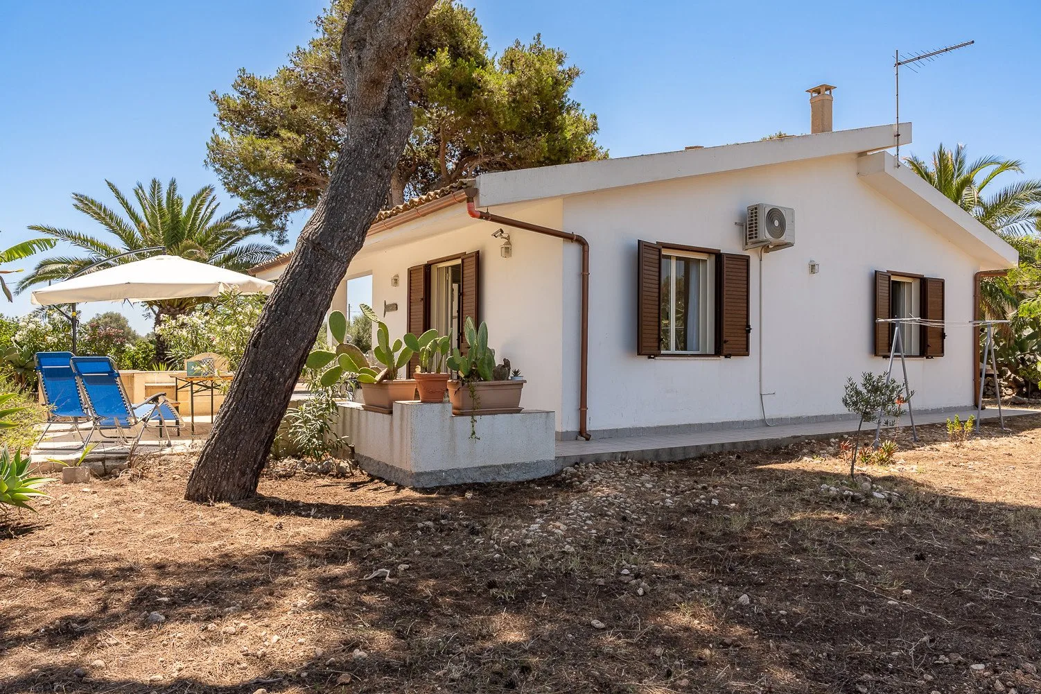 Una casa blanca está junto a un patio sombreado con asientos azules al aire libre.