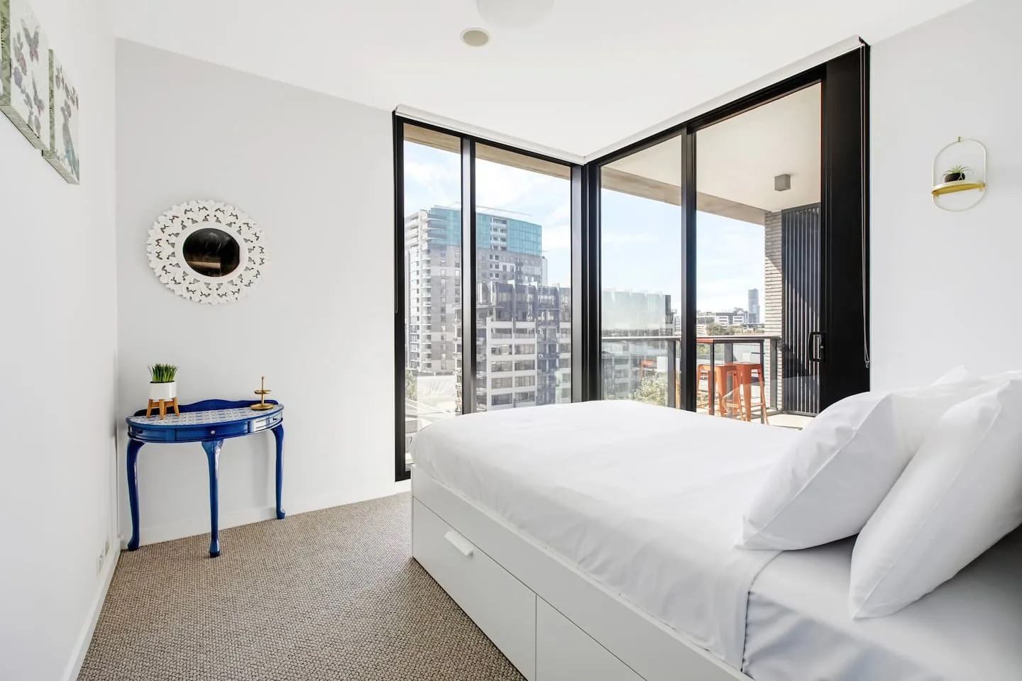 A bedroom with a bed beside large windows overlooking nearby buildings.