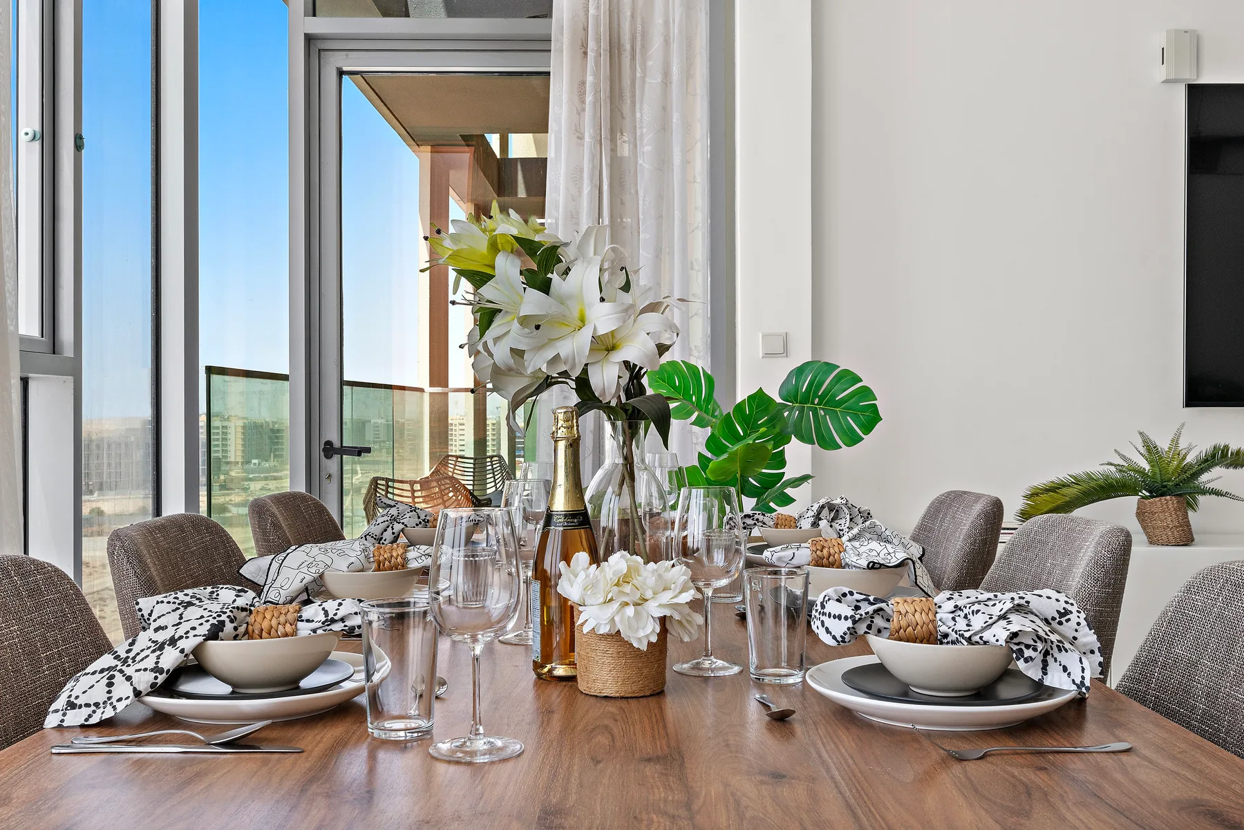 Una mesa de comedor está puesta con platos y flores junto a ventanales.
