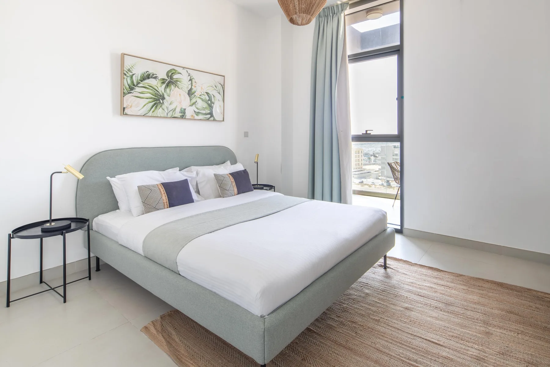 Un dormitorio luminoso con una cama tendida, arte botánico en la pared y una ventana alta.