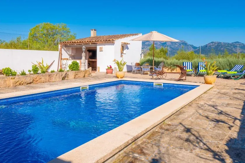 A swimming pool beside a white house, with loungers and an umbrella on the patio.