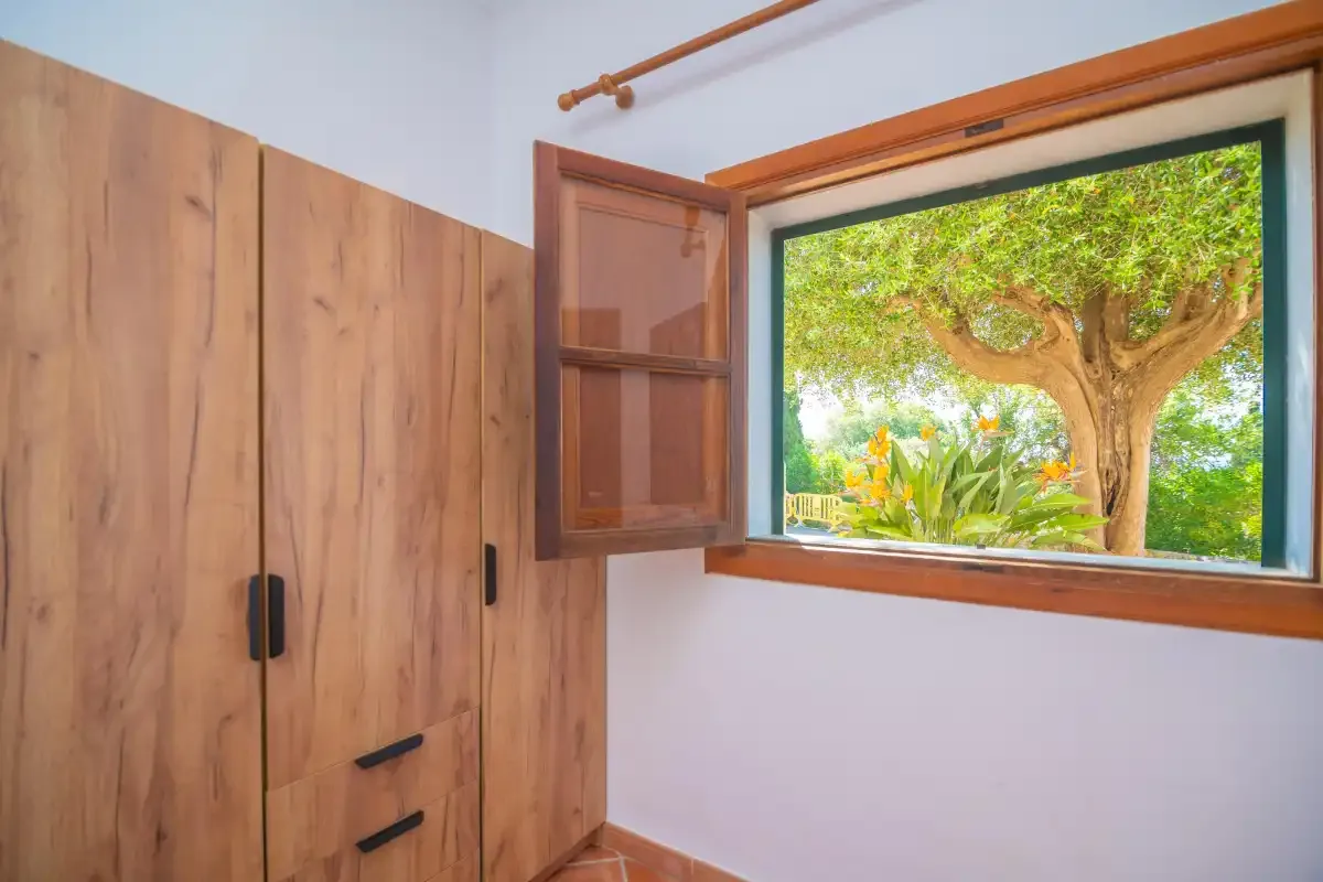 A bedroom window opens onto a leafy garden beside a wooden wardrobe.