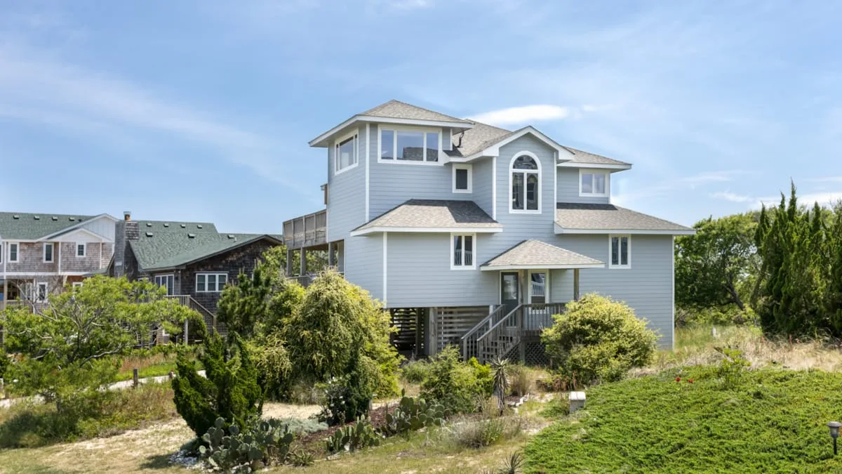 A raised coastal-style house stands among greenery beneath a clear sky.