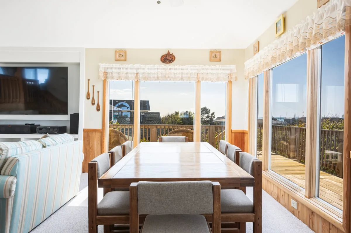 A bright dining area with a long table beside large windows opening onto a deck.
