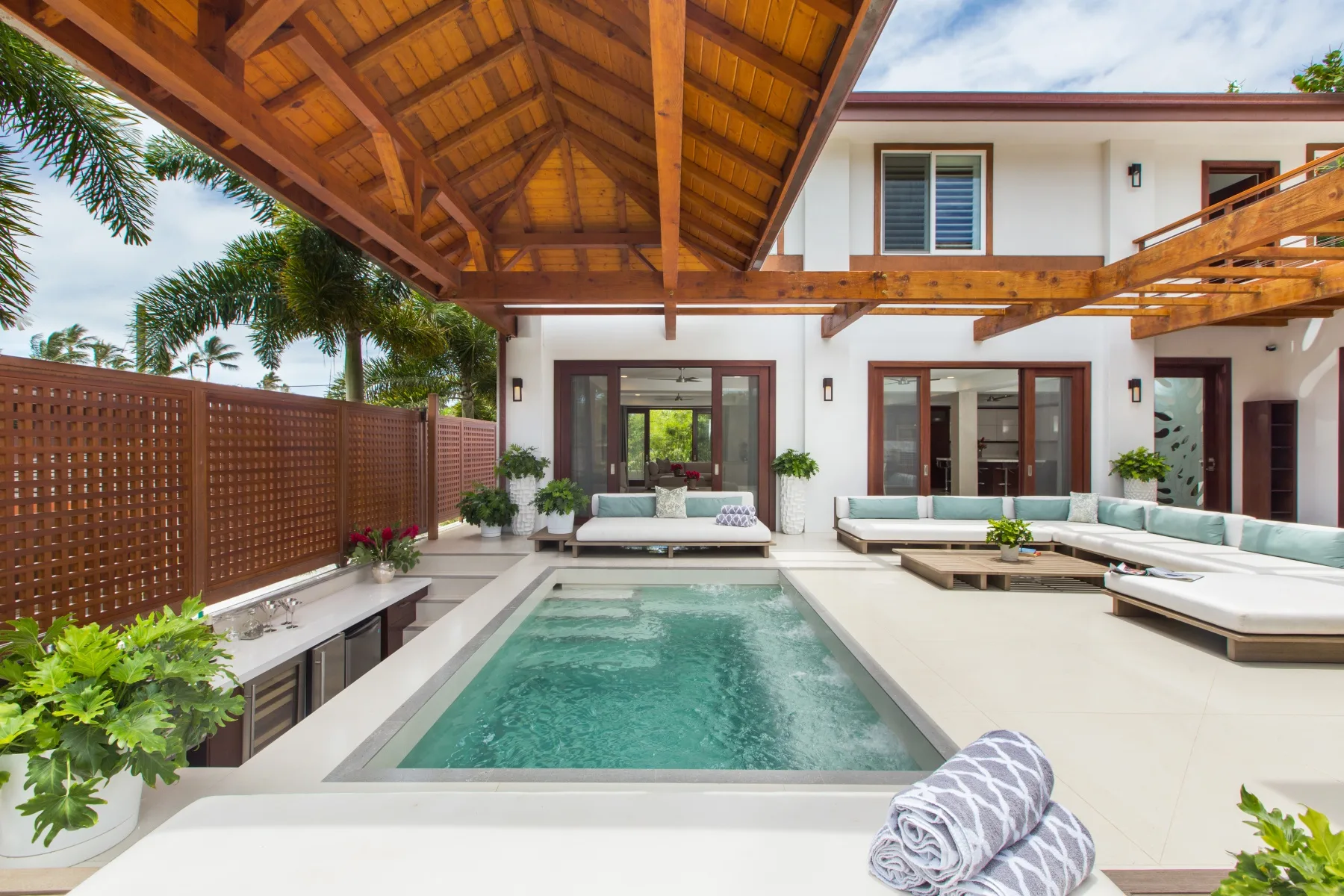 Una piscina en un patio está bajo un techo de madera junto a un edificio blanco.