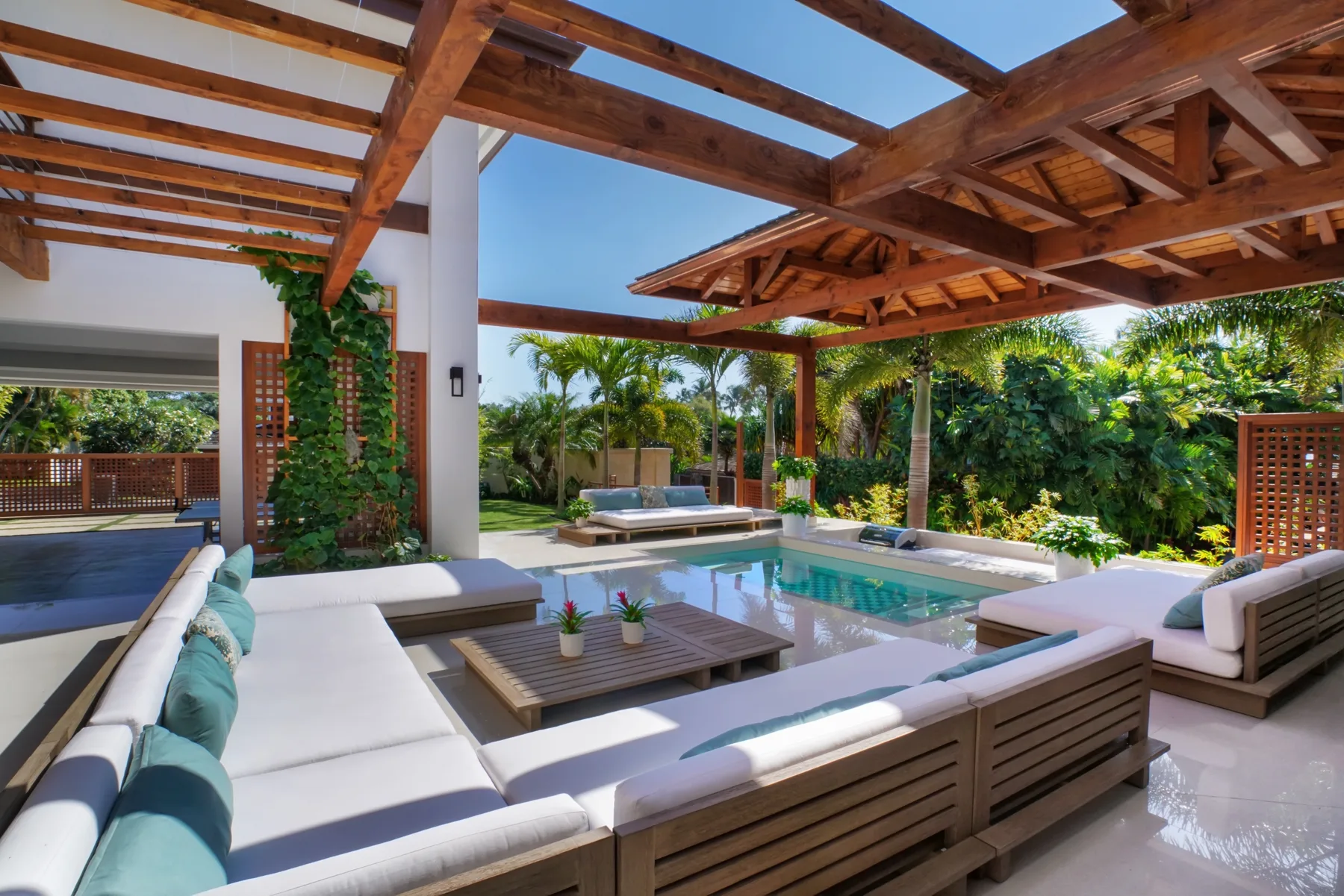 Una sala de estar al aire libre con asientos acolchados y una piscina bajo una pérgola de madera.