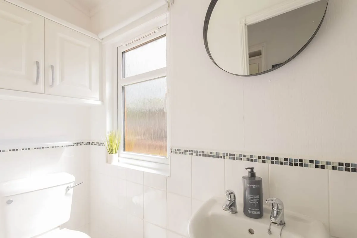 Un baño luminoso con azulejos, una ventana, un lavabo y un espejo redondo.
