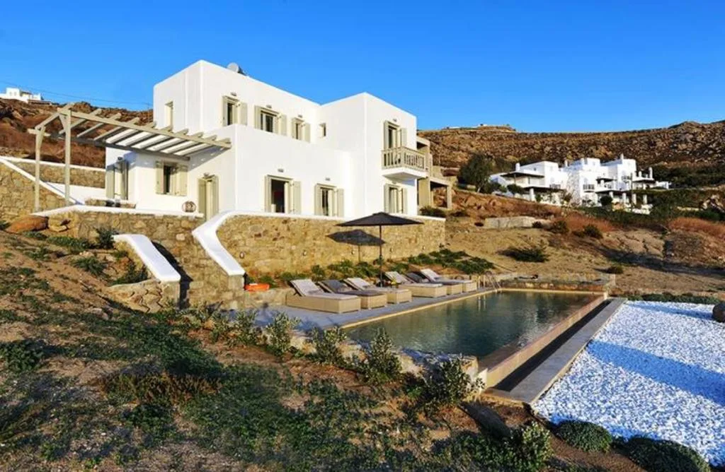 Una villa blanca da a una piscina y tumbonas en una ladera.