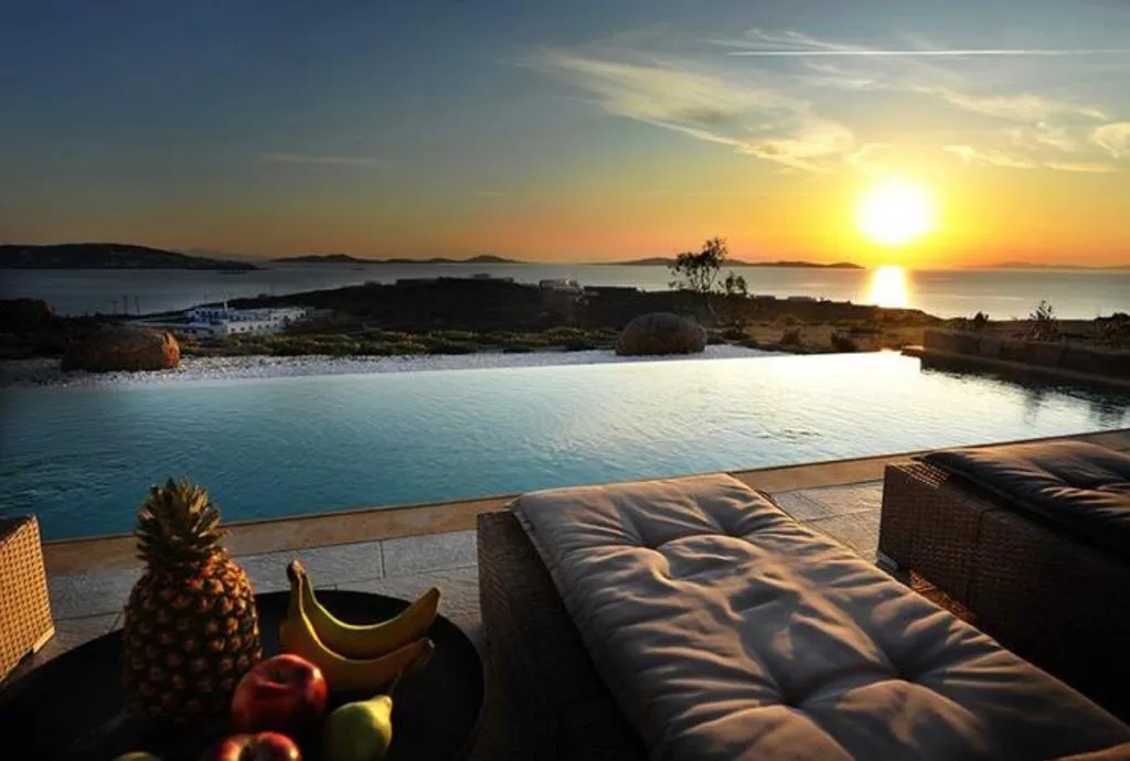 Una piscina infinita mira al mar al atardecer, con tumbonas acolchadas al lado.