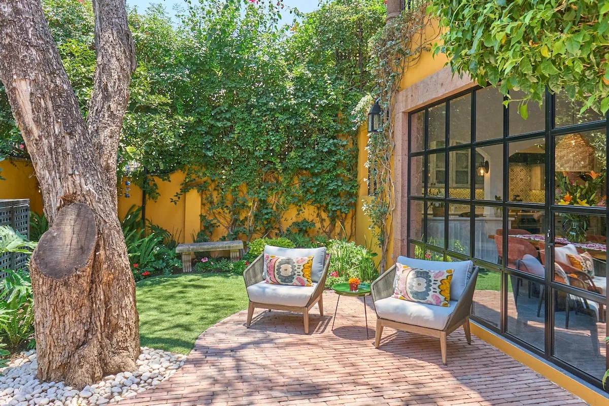 A leafy courtyard patio has two cushioned seats beside large glass windows.