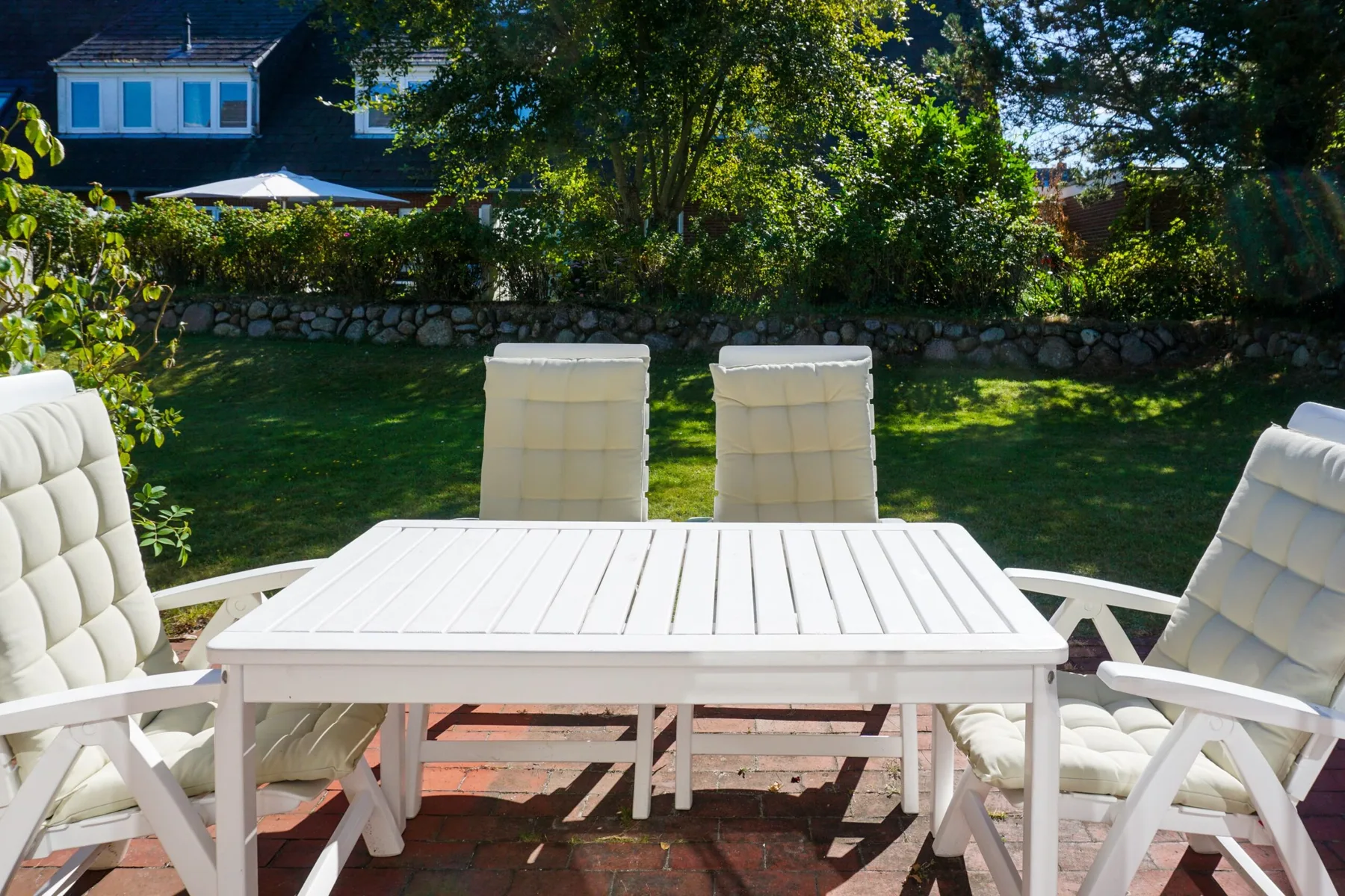 Una mesa blanca de exterior y asientos acolchados están en un patio de ladrillo junto a un jardín.