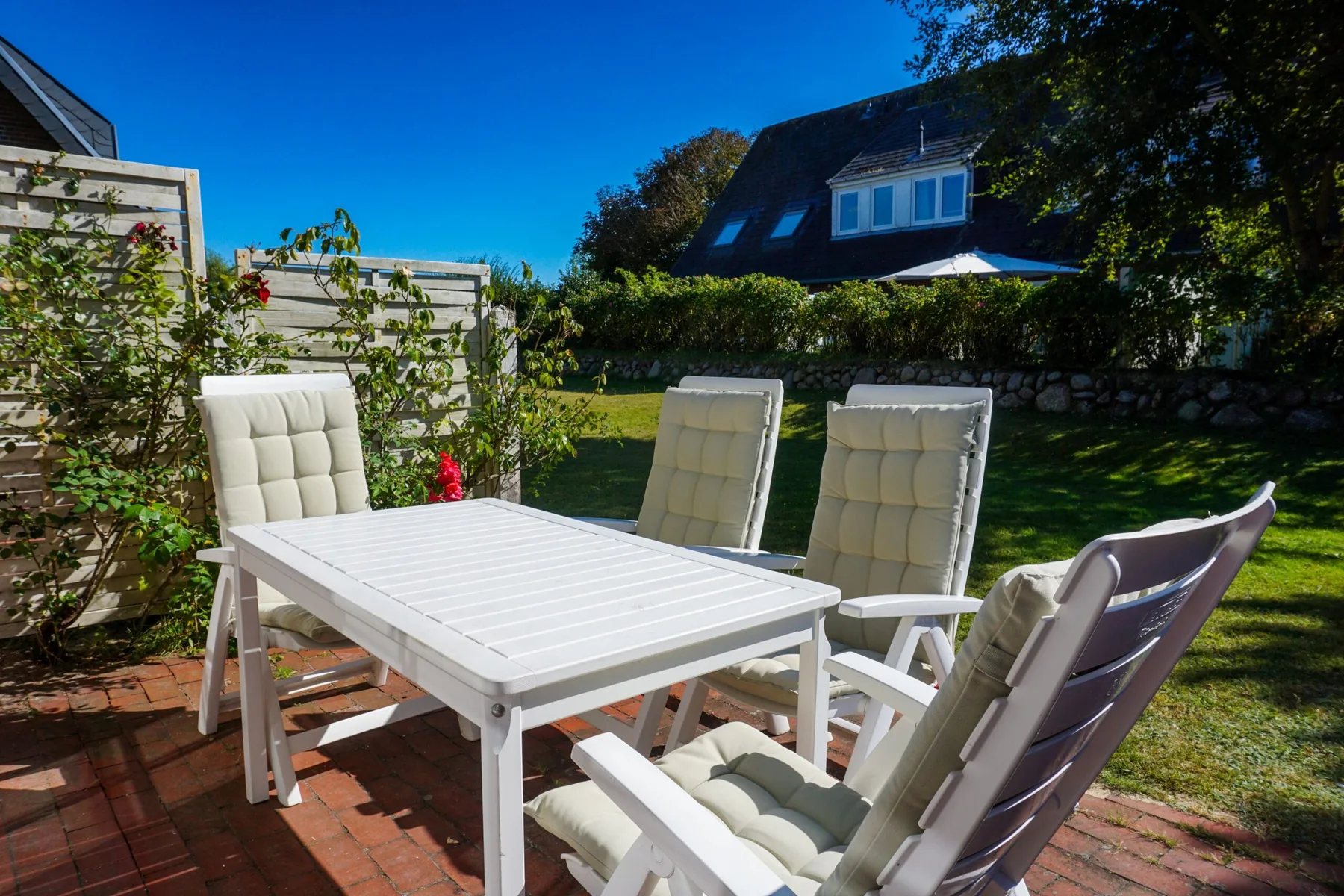 Un patio exterior tiene una mesa blanca y asientos acolchados junto a un jardín.