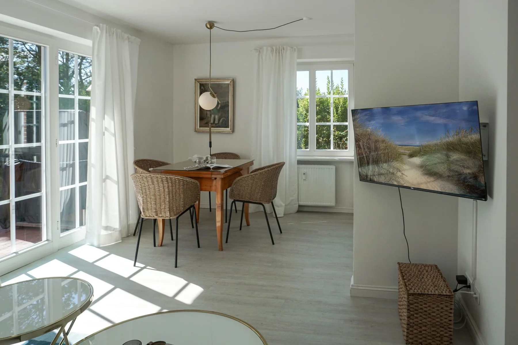 Una luminosa zona de comedor y estar con mesa de madera, asientos tejidos y grandes ventanas.