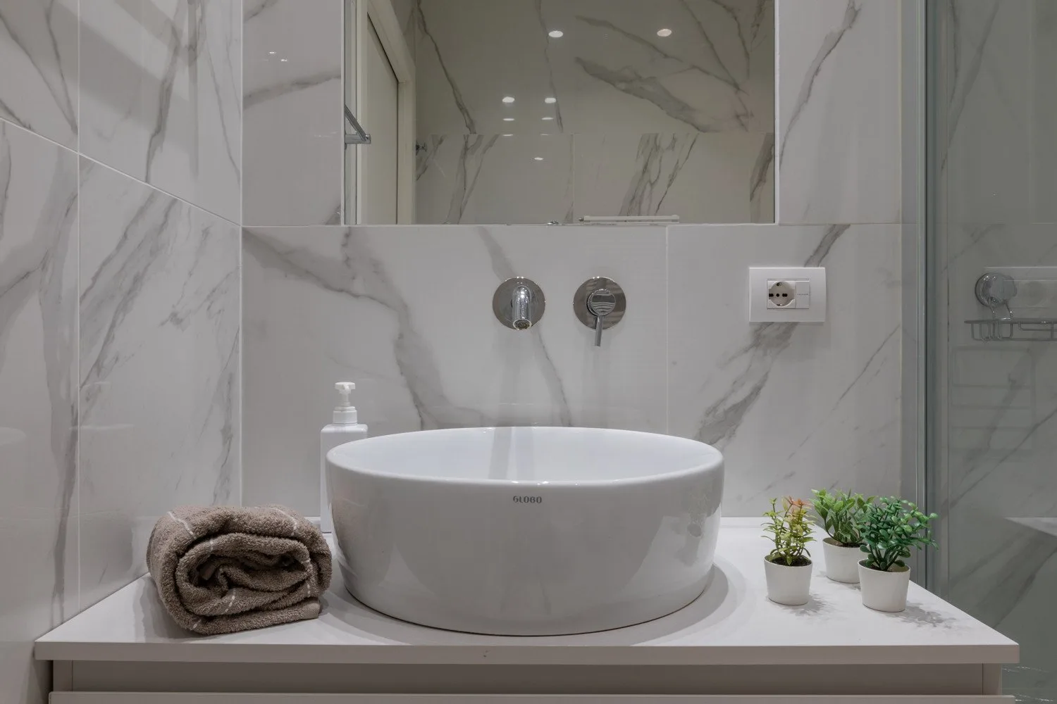 Un tocador de baño moderno con lavabo sobre encimera blanco y paredes con diseño de mármol.