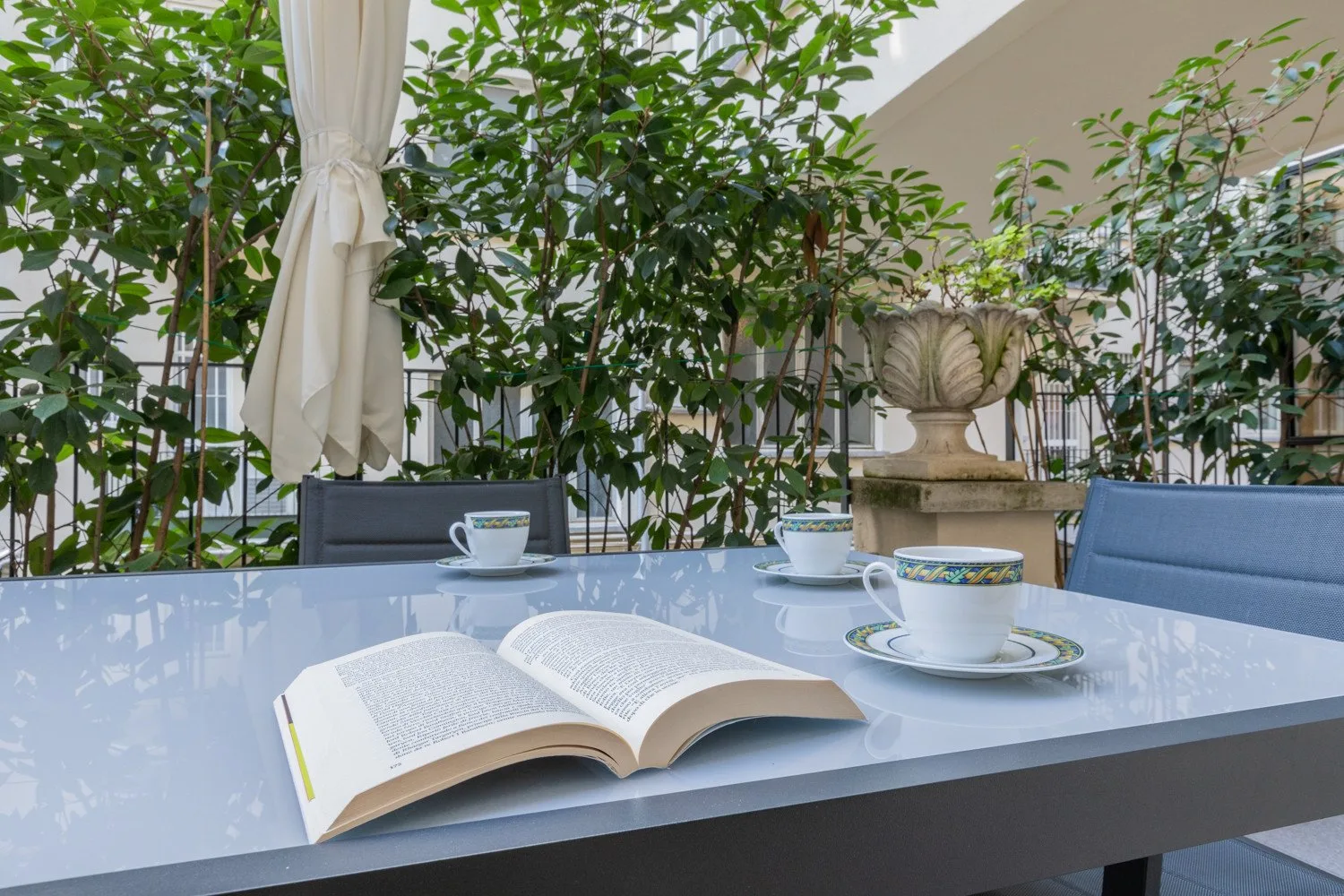 Una mesa de patio con un libro abierto, tazas y plantas frondosas.
