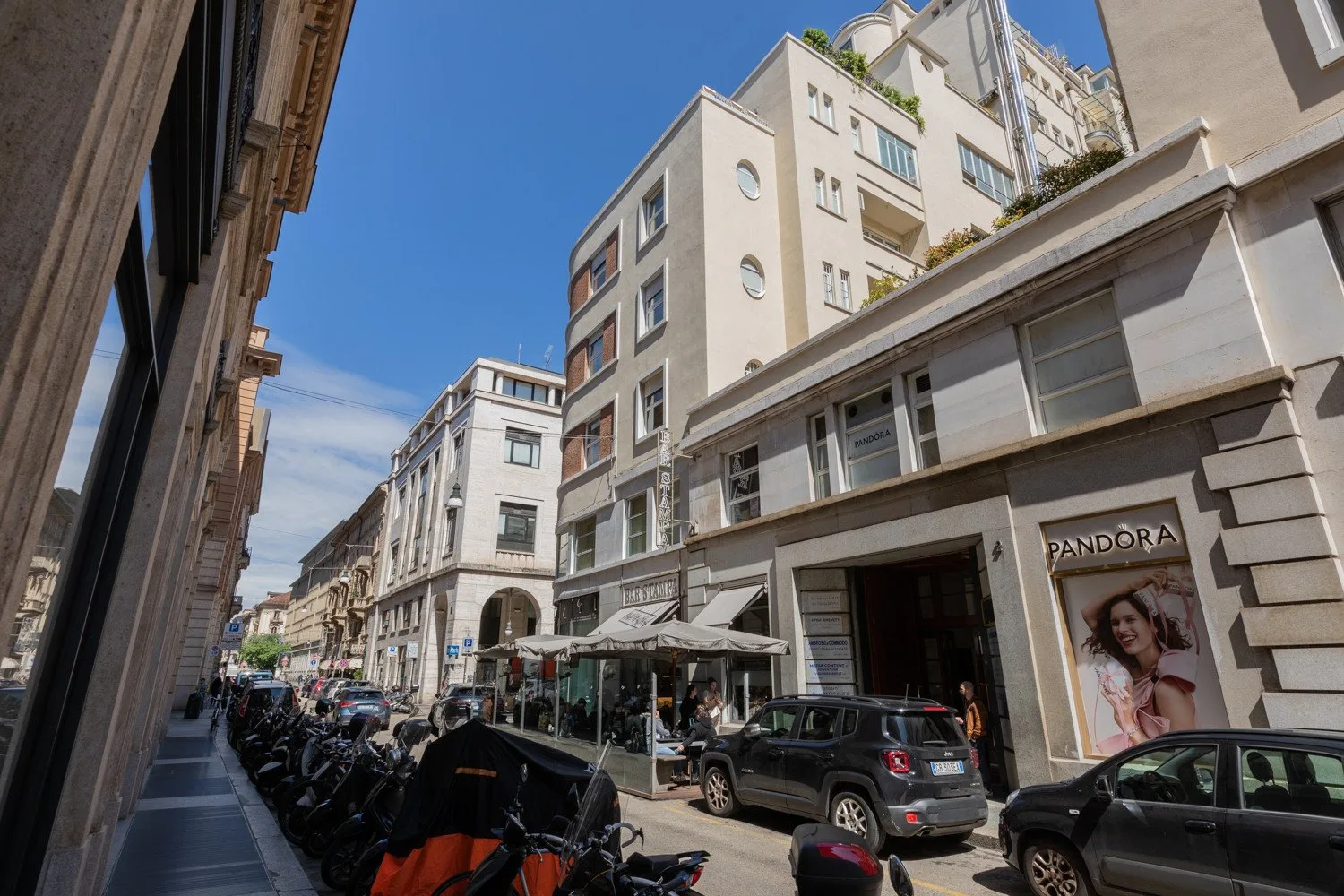 Una calle urbana con edificios de varias plantas, vehículos estacionados y mesas de café en la acera.