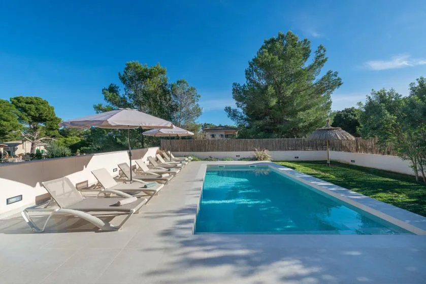 Una piscina junto a un patio pavimentado con tumbonas y una sombrilla.