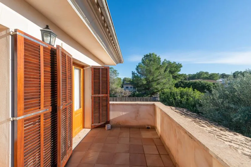 Un balcón embaldosado con contraventanas de madera da a árboles y vegetación.