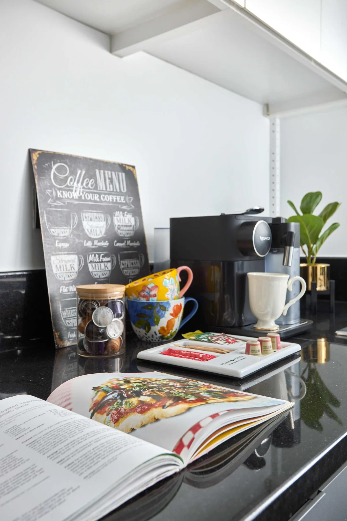 Una estación de café con una cafetera espresso, tazas coloridas y revistas abiertas sobre la encimera.