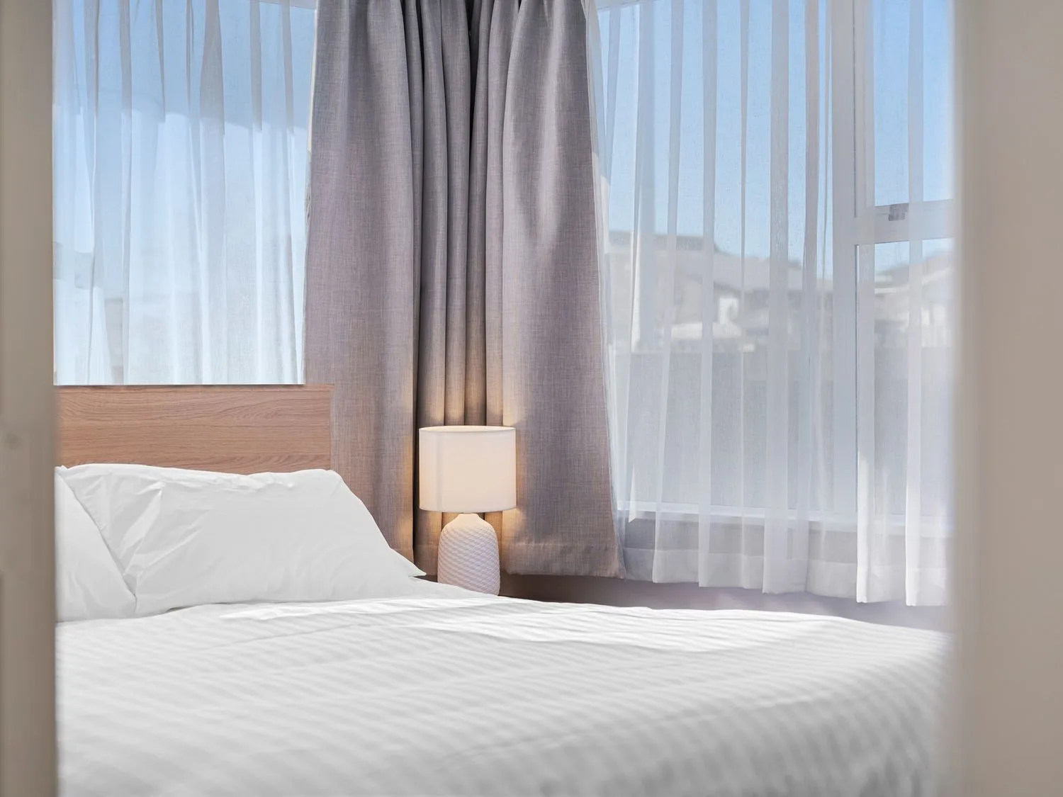 A bedroom with a bed having white bedding a wooden headboard a lit lamp and windows with light grey and sheer curtains at Owhata Retreat - Rotorua Holiday Home in Rotorua