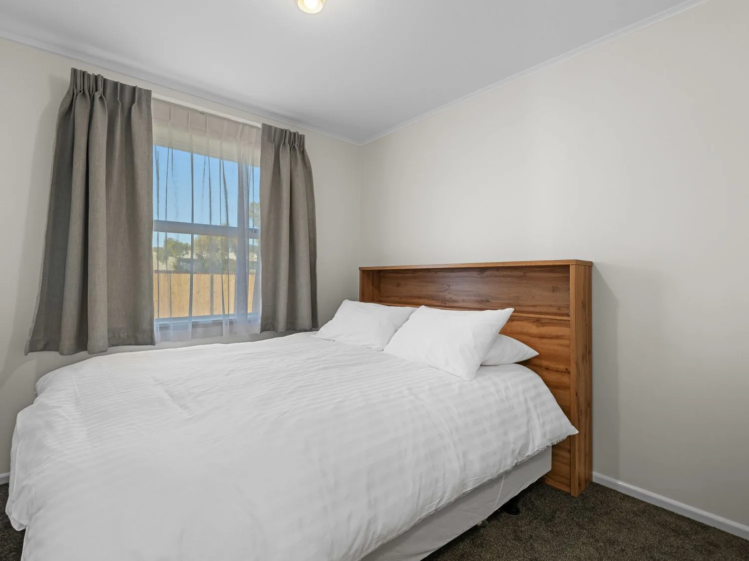 A bedroom with a bed with white bedding and wooden headboard next to a window with curtains at Owhata Retreat - Rotorua Holiday Home in Rotorua