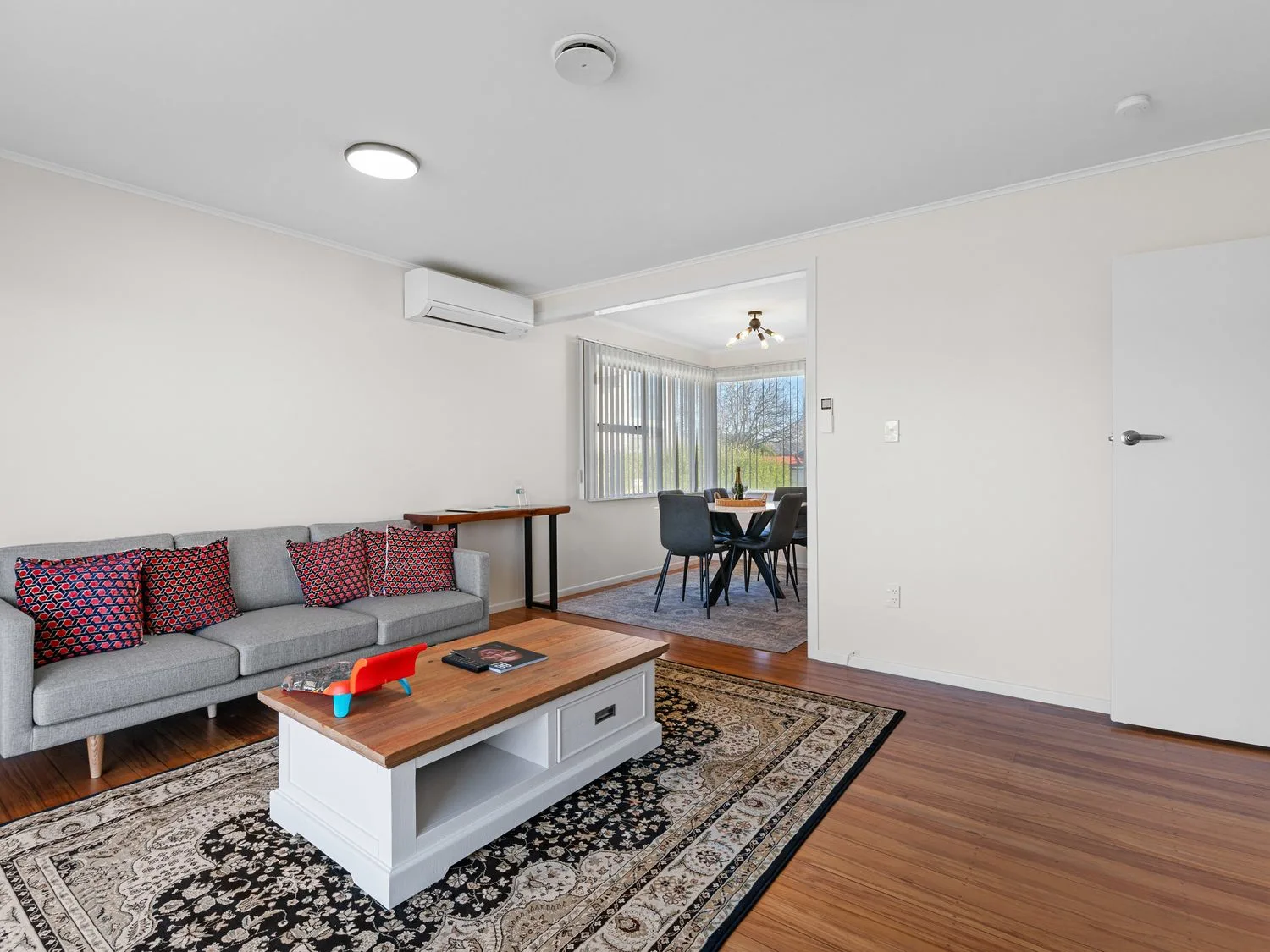 A living room with a gray sofa and patterned cushions a white coffee table on a rug and a dining area with chairs and a window in Owhata Retreat - Rotorua Holiday Home in Rotorua