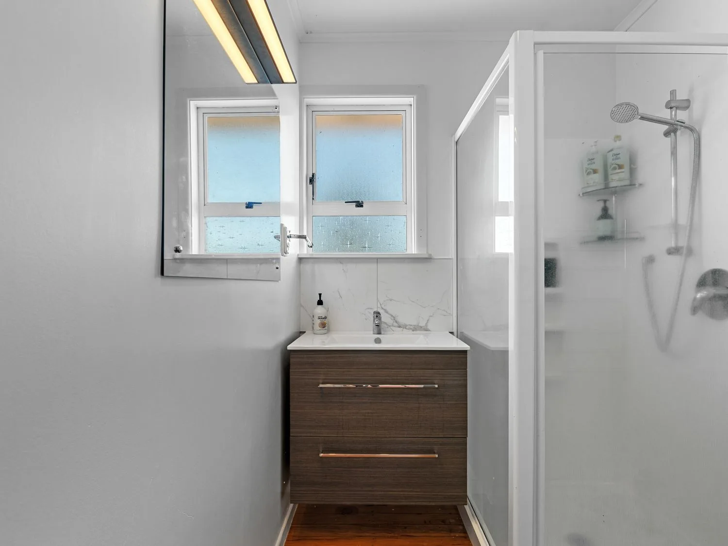 A bathroom with a vanity and sink beneath two windows beside a glass shower.