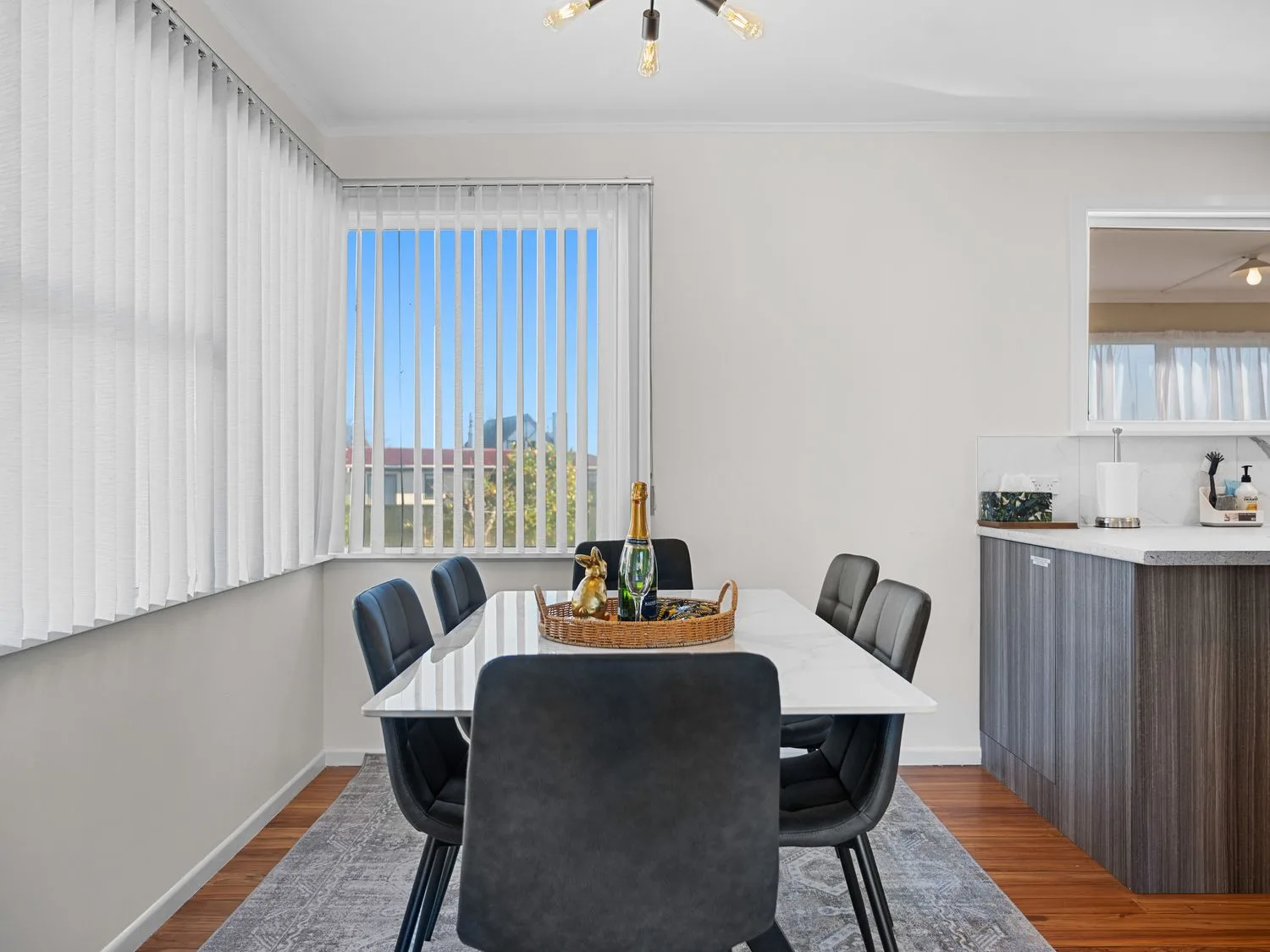 A dining area with a white table surrounded by six black chairs and a large window with vertical blinds at Owhata Retreat - Rotorua Holiday Home in Rotorua