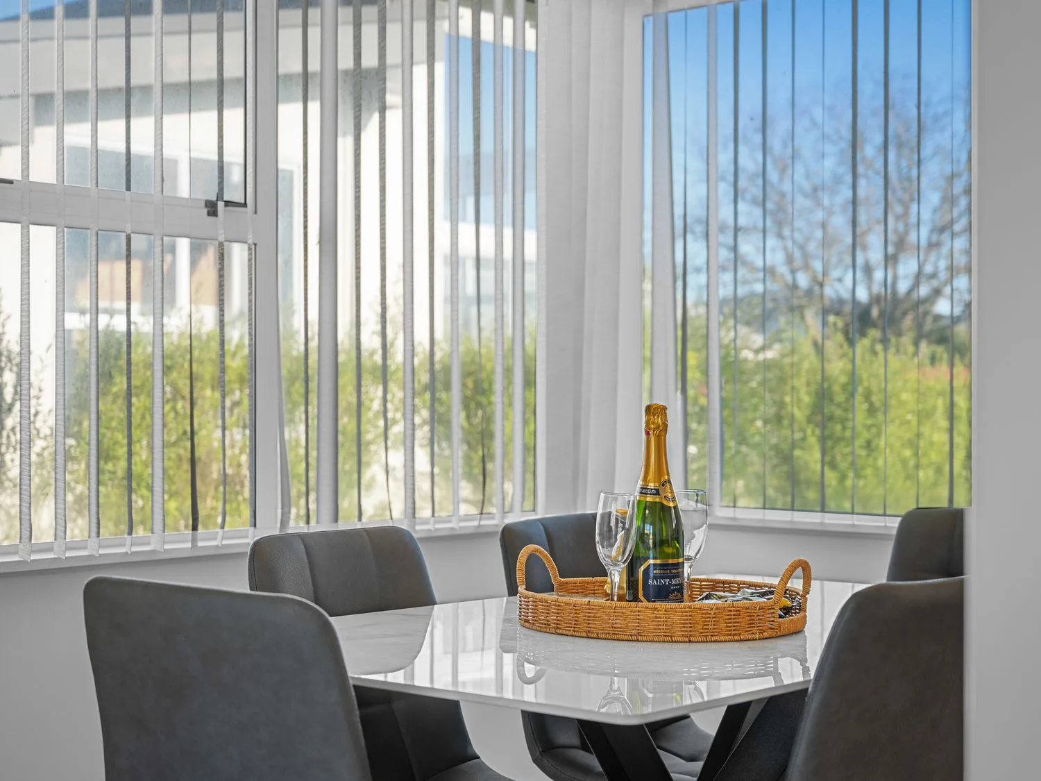 A dining area with a table and seating beside large windows with vertical blinds.