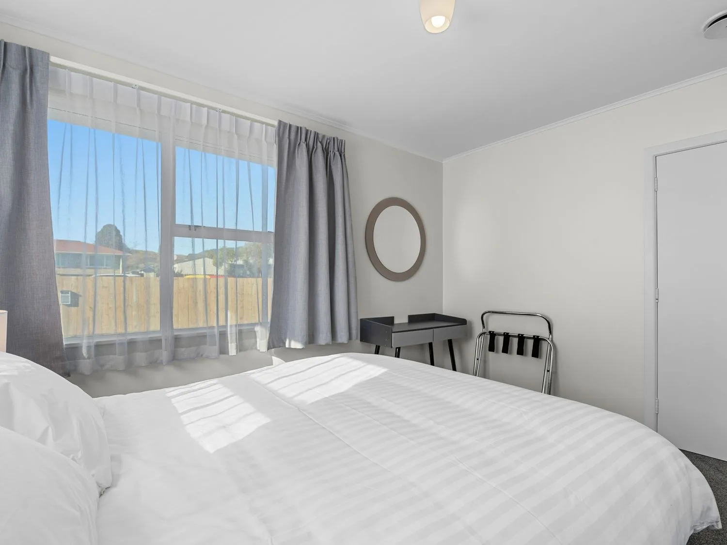 A bedroom with a bed near a window with gray curtains a small black desk a round mirror on the wall and a luggage rack at Owhata Retreat - Rotorua Holiday Home Rotorua
